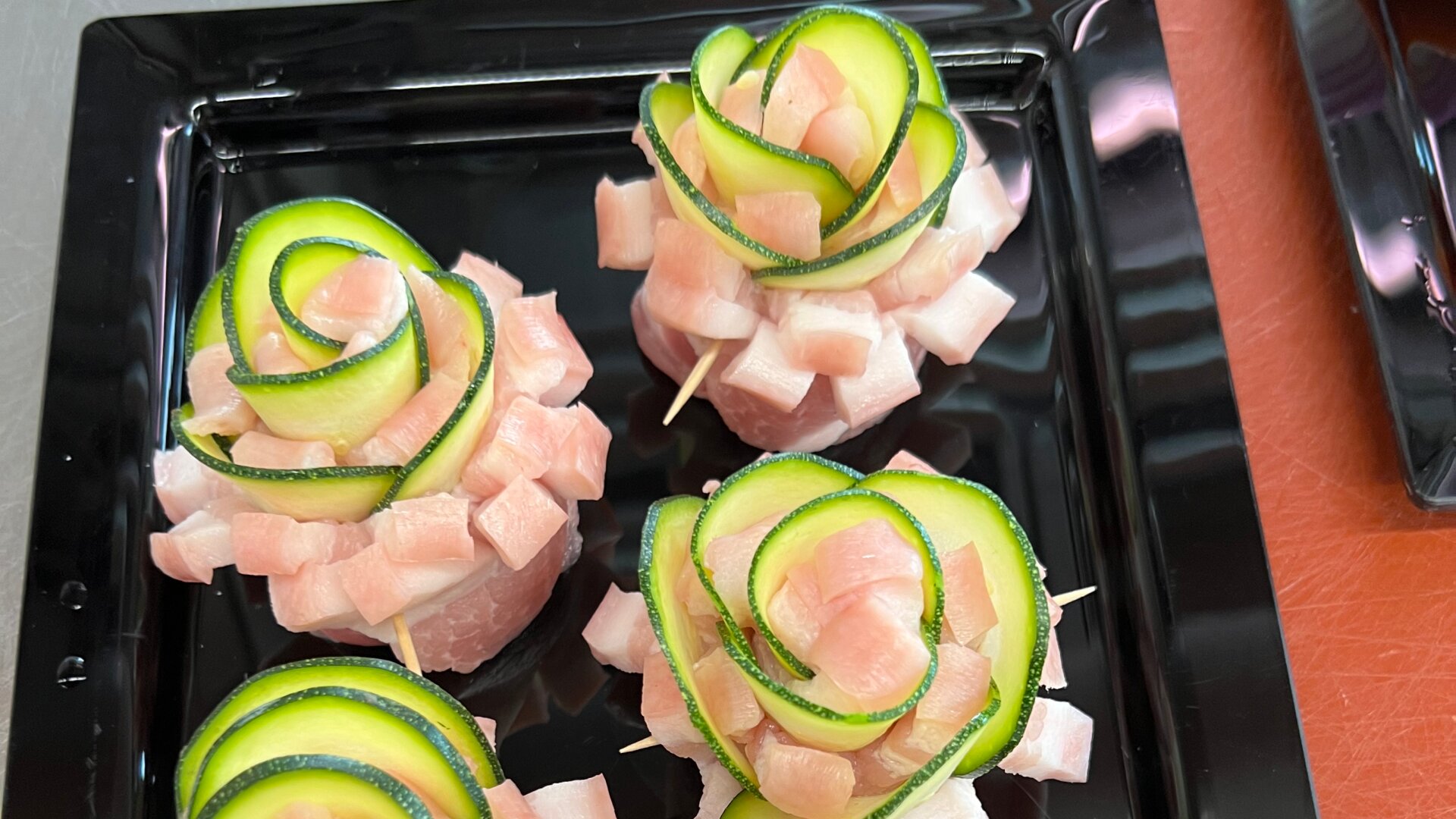 Vier dekorative Sushi-Stücke aus rohen Fischwürfeln und dünnen Gurkenscheiben, die auf einem schwarzen Tablett in Form einer Rose angeordnet sind.