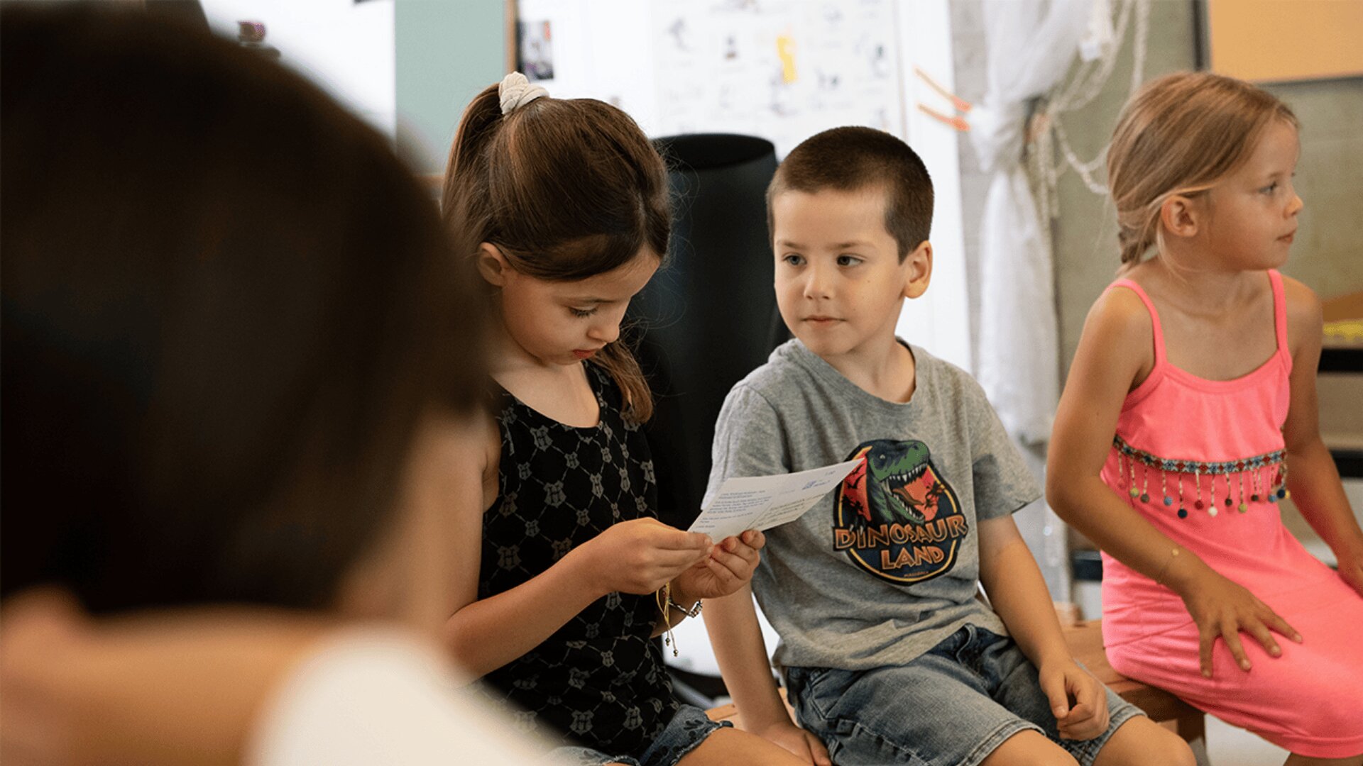 Drei kleine Kinder sitzen in einem Haus. Ein Mädchen liest von einem Blatt Papier ab, während ein Junge, der ein Dinosaurier-T-Shirt trägt, sie ansieht. Ein anderes Mädchen in einem rosa Kleid sitzt daneben und schaut weg.