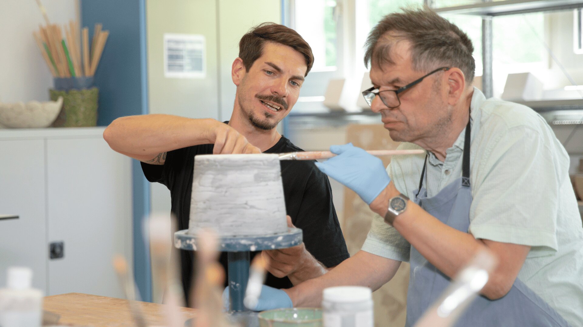 Zwei Männer arbeiten gemeinsam an einem Töpferprojekt. Ein Mann formt einen Tontopf auf einem Spinnrad, während der andere, der Handschuhe und eine Schürze trägt, ein Holzwerkzeug hält, um den Topf zu glätten. Im Vordergrund sind Kunstgegenstände zu sehen.