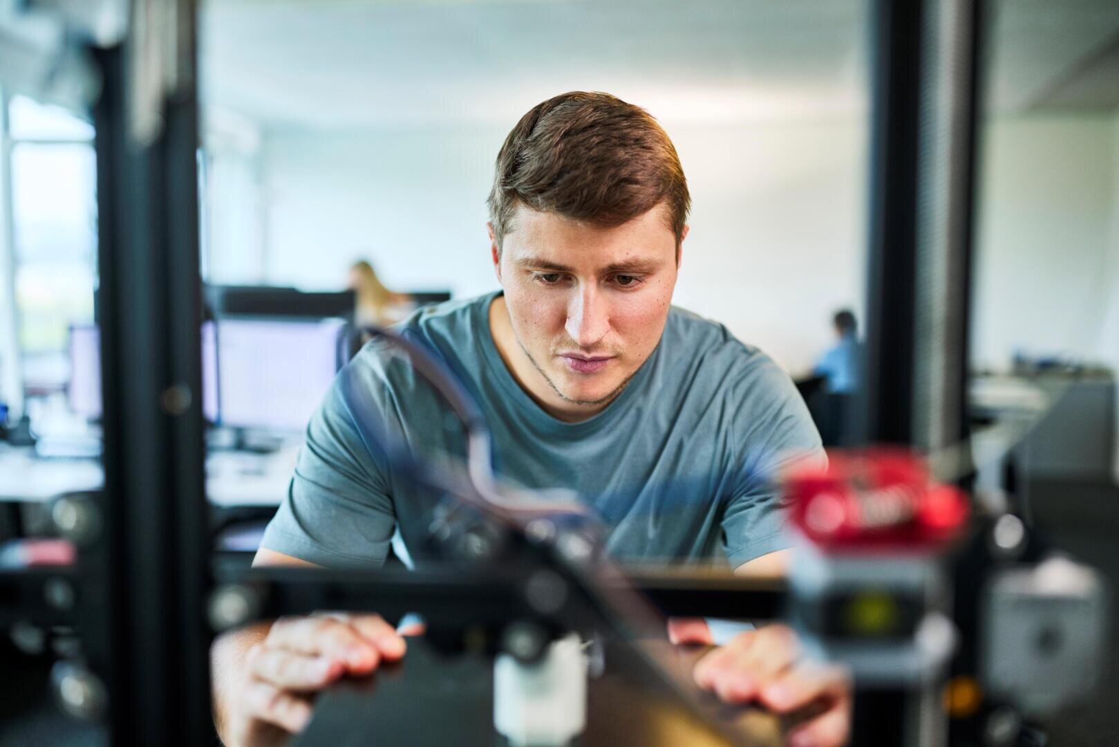 Eine Person konzentriert sich intensiv auf die Bedienung oder Überwachung eines 3D-Druckers in einem Büro oder Labor, wobei im unscharfen Hintergrund Computerbildschirme und eine weitere Person zu sehen sind.