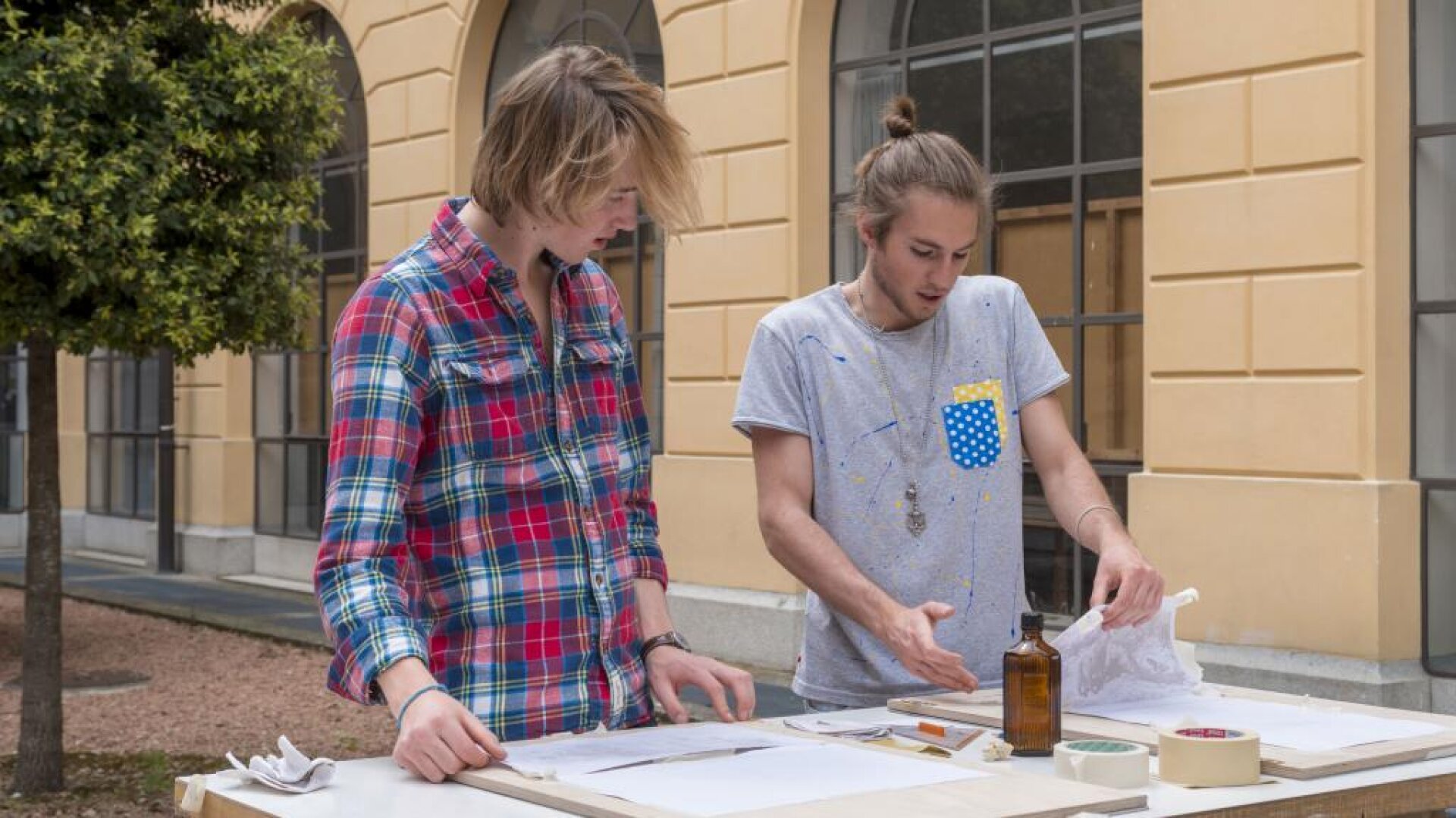 Zwei junge Leute arbeiten an einem Tisch im Freien, der mit Papieren, Klebeband und einer Flasche bedeckt ist. Einer trägt ein kariertes Hemd und der andere ein graues T-Shirt mit Farbflecken. Sie scheinen sich auf ein Kunst- oder Handwerksprojekt zu konzentrieren.