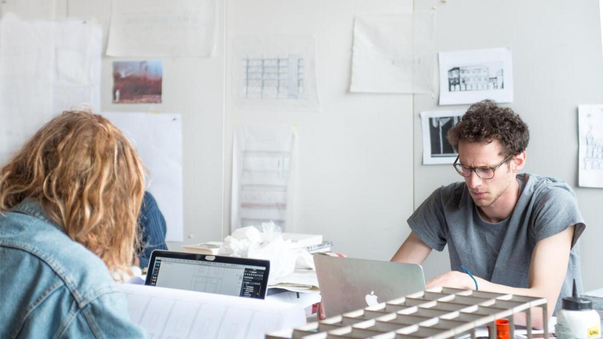 Zwei Personen arbeiten an Schreibtischen mit Laptops in einem hellen Raum, umgeben von Architekturzeichnungen und Modellen. Papiere und Skizzen sind an die weißen Wände hinter ihnen gepinnt. Die Atmosphäre wirkt konzentriert und kreativ.