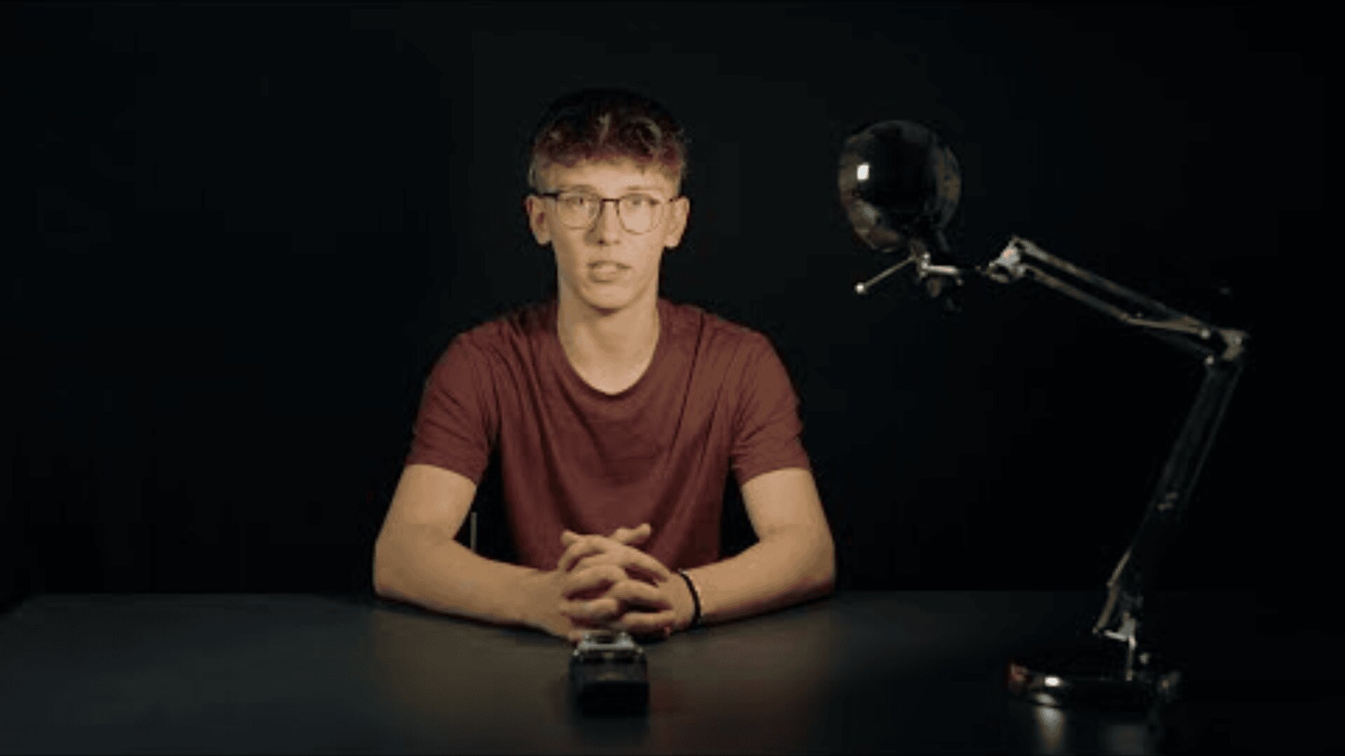 Eine junge Person mit kurzen Haaren und Brille sitzt an einem Schreibtisch in einem dunklen Raum, die Hände gefaltet und der Kamera zugewandt. Rechts steht eine moderne Schreibtischlampe, die Licht auf den Tisch wirft.