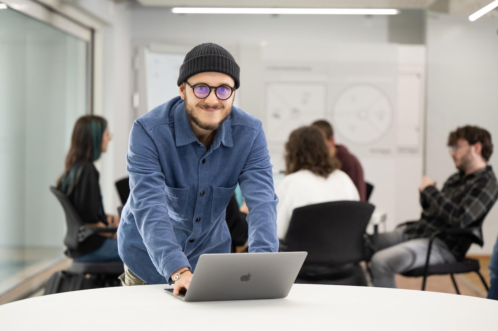 Ein Mann in einem blauen Hemd und einer schwarzen Mütze lächelt, während er sich mit einem Laptop an einen runden Tisch lehnt. Im Hintergrund sitzen vier Personen und unterhalten sich in einem modernen, hellen Büroraum.