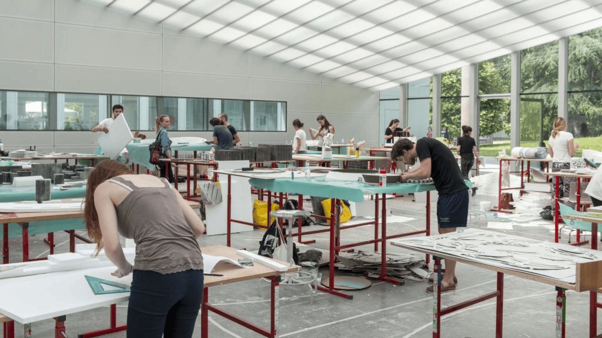 Die Schüler arbeiten an Tischen in einem geräumigen, gut beleuchteten Atelier mit hohen Decken und großen Fenstern an großen Kunst- oder Designprojekten. Der Raum ist mit Materialien, Papier und kreativen Aktivitäten gefüllt.