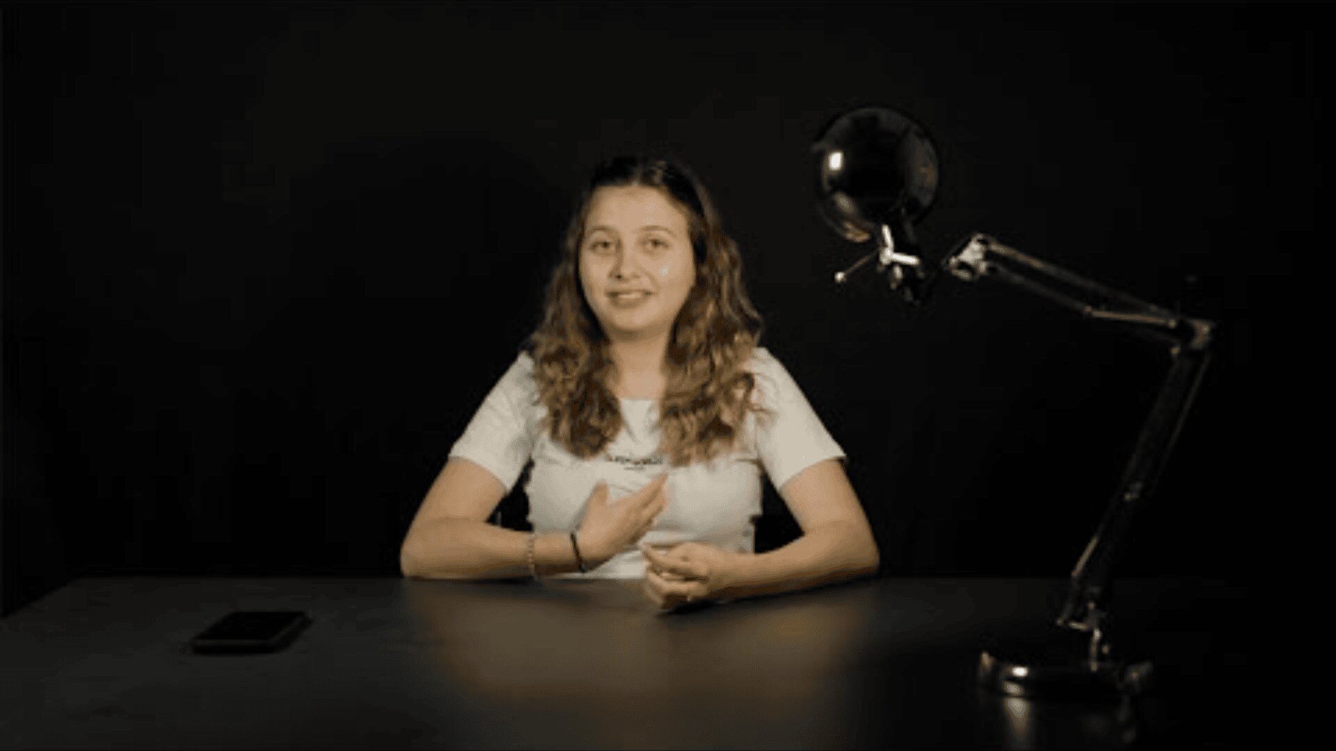 Eine Frau mit langem, gewelltem Haar sitzt an einem dunklen Tisch vor einem schwarzen Hintergrund. Sie trägt ein weißes Hemd und gestikuliert mit ihren Händen. Eine Schreibtischlampe und ein Telefon stehen ebenfalls auf dem Tisch.