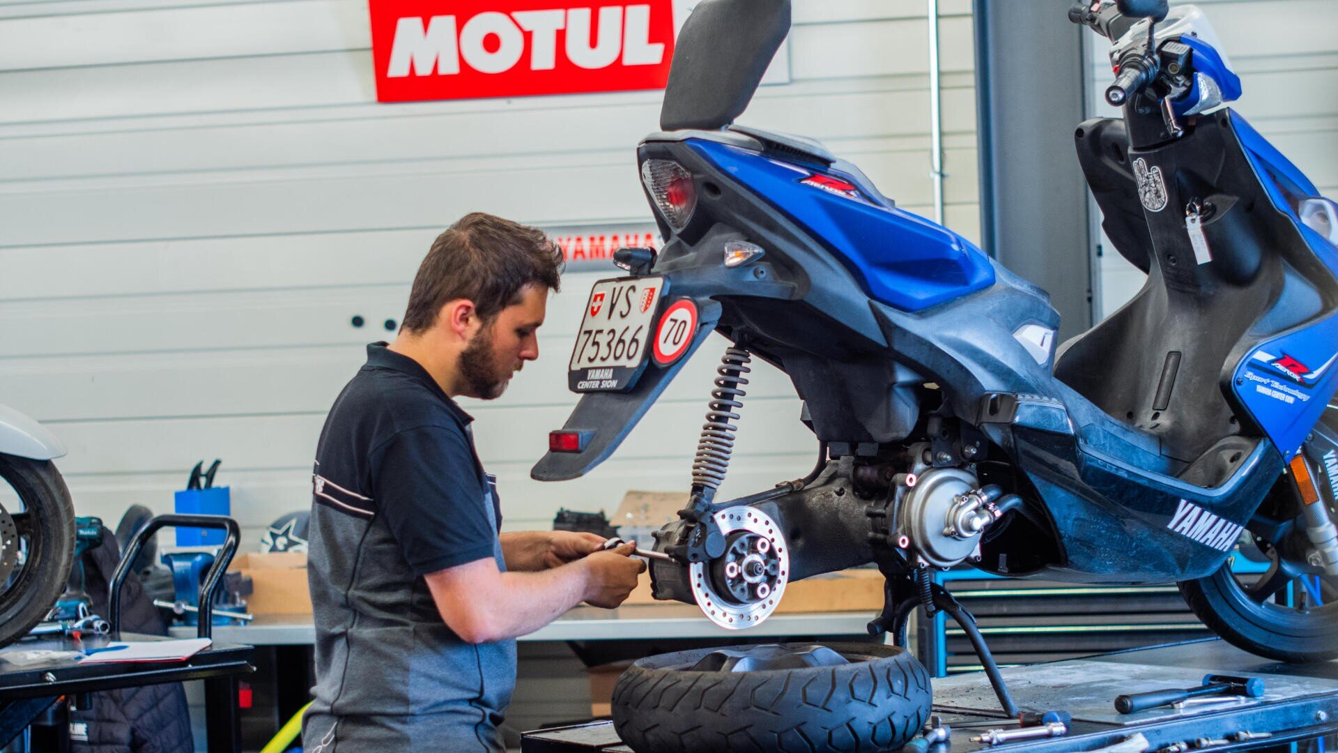 Ein Mechaniker arbeitet an einem blauen Yamaha-Roller mit ausgebautem Hinterrad in einer Werkstatt. Werkzeuge und Ausrüstung sind zu sehen, und im Hintergrund hängt ein Motul-Schild an der Wand.