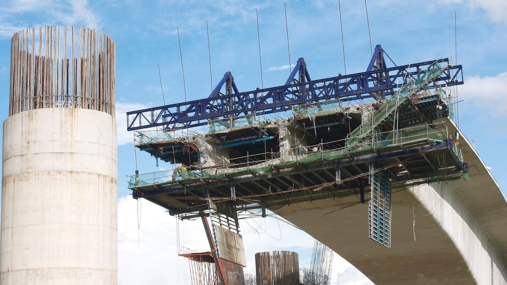 Eine im Bau befindliche Betonbrücke mit freiliegenden Bewehrungsstützen und Gerüsten, einer teilweise fertiggestellten Spannweite und blauen Stützbalken vor einem teilweise bewölkten Himmel.