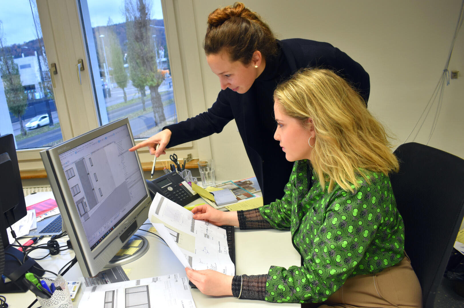 Zwei Frauen arbeiten gemeinsam an einem Schreibtisch; die eine sitzt, hält Dokumente in der Hand und schaut auf einen Computerbildschirm, während die andere neben ihr steht und auf den Bildschirm zeigt, beide konzentriert auf Architekturpläne.