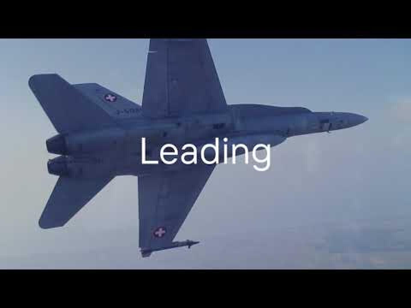 Ein militärischer Kampfjet mit Schweizer Insignien fliegt am Himmel. Das Wort "Leading" erscheint in großer weißer Schrift in der Mitte des Bildes.