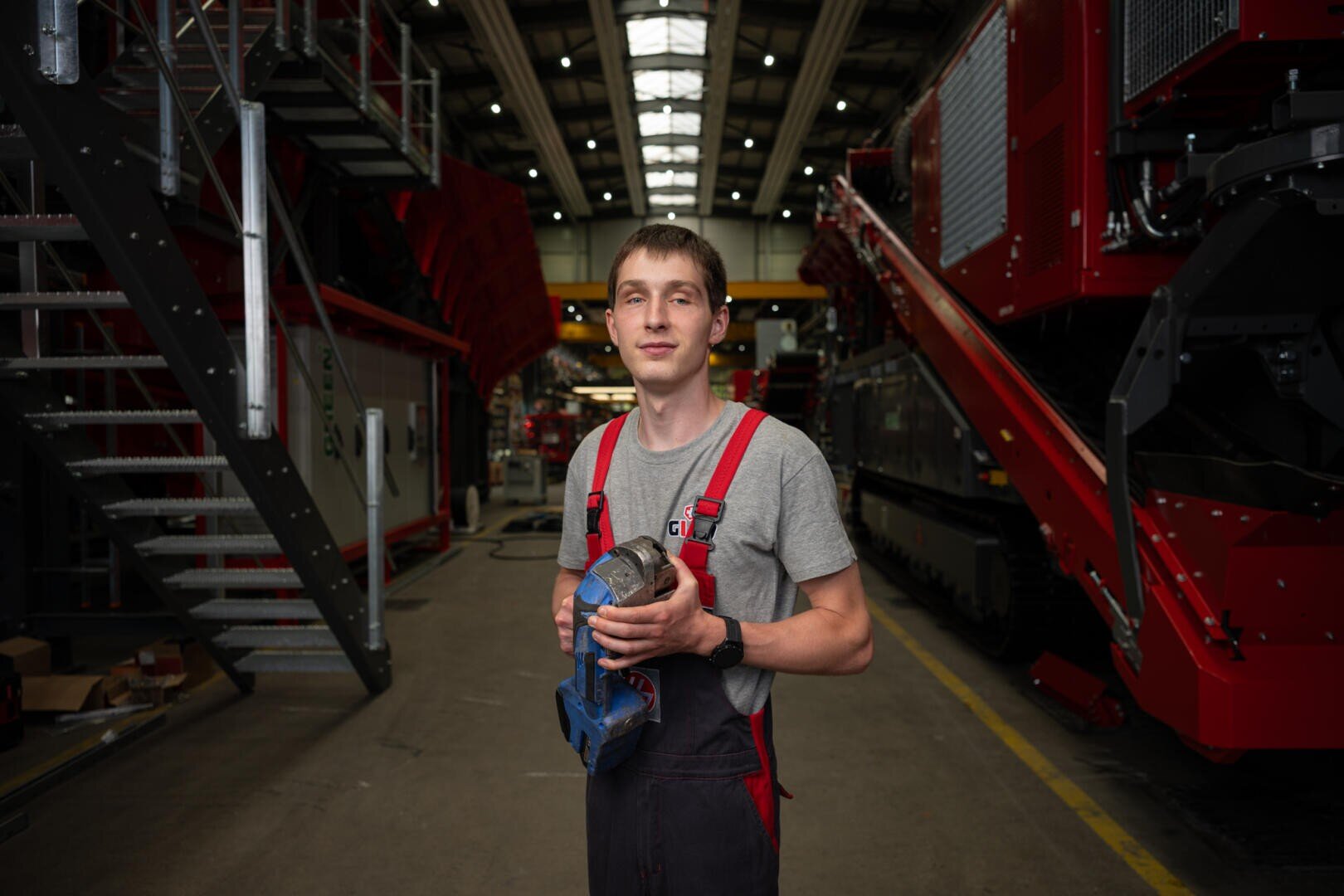 Ein junger Mann in Latzhose und einem grauen Hemd steht in einer Fabrik, ein blaues Elektrowerkzeug in der Hand, umgeben von großen roten Industriemaschinen und -anlagen.