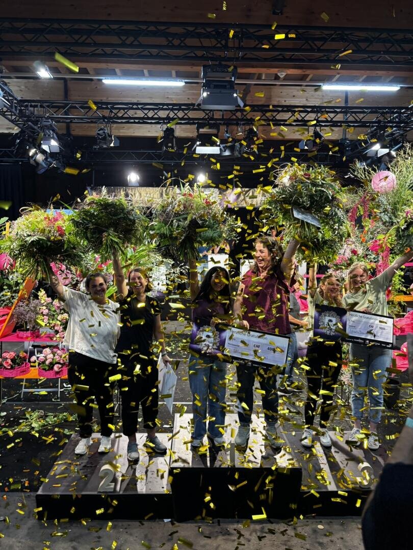 Sechs Personen stehen auf Podesten, halten Blumen und Urkunden in den Händen und sind von Konfetti umgeben. Bunte Blumenarrangements schmücken die Bühne, über der helle Beleuchtung und Holzbalken zu sehen sind. Festliche Atmosphäre in einem Innenraum.