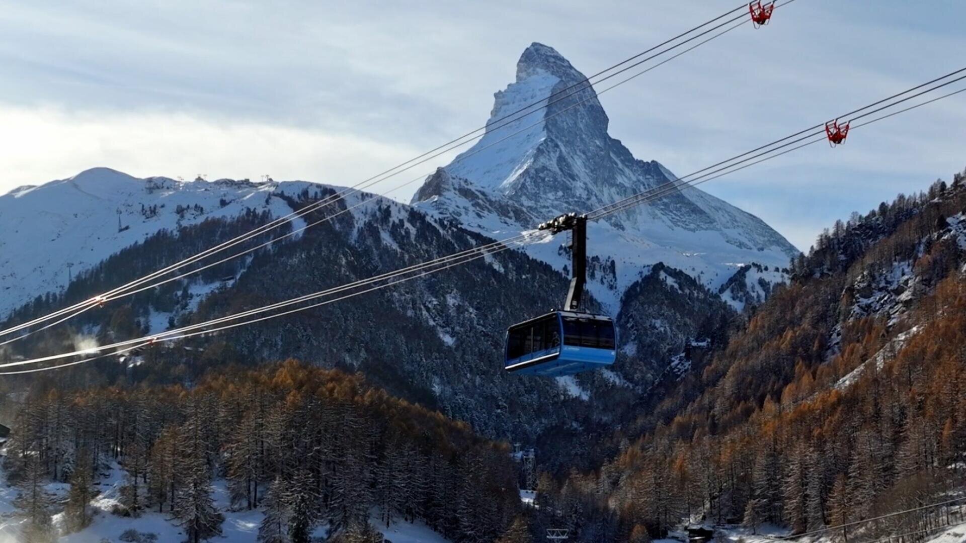 Eine Seilbahn fährt über ein verschneites, bewaldetes Tal hinweg in Richtung des Matterhorns.