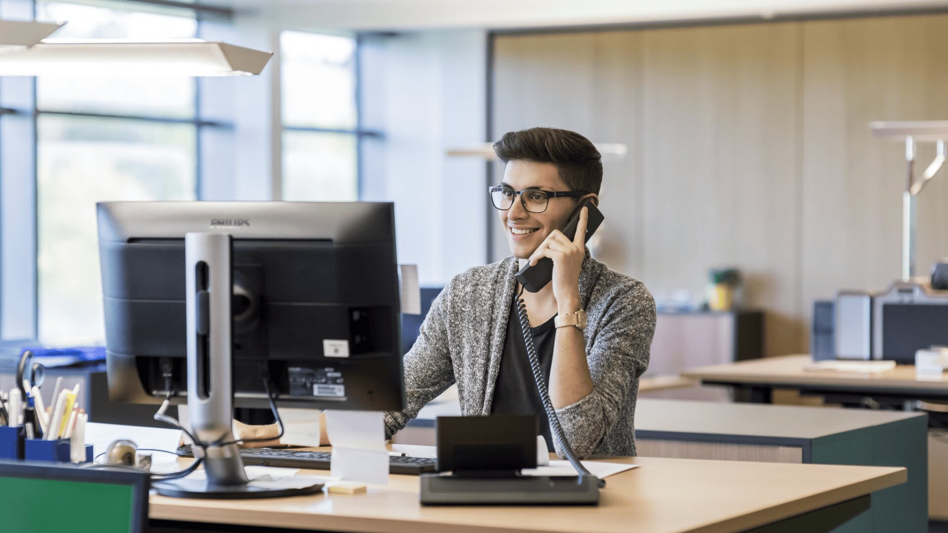 Ein junger Mann mit Brille sitzt an einem Schreibtisch in einem modernen Büro und lächelt, während er telefoniert und auf einen Computerbildschirm schaut. Das Büro hat große Fenster und eine helle, offene Gestaltung.