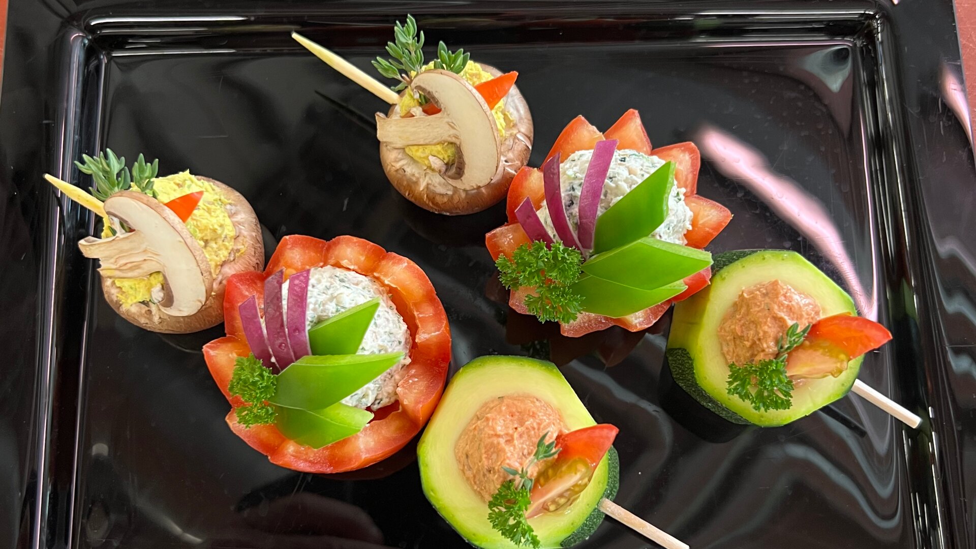 Eine Auswahl bunter Vorspeisen auf einem schwarzen Tablett, darunter gefüllte Pilze, Tomaten und Avocados, garniert mit Kräutern, geschnittenem Gemüse und Spießen zur Dekoration.