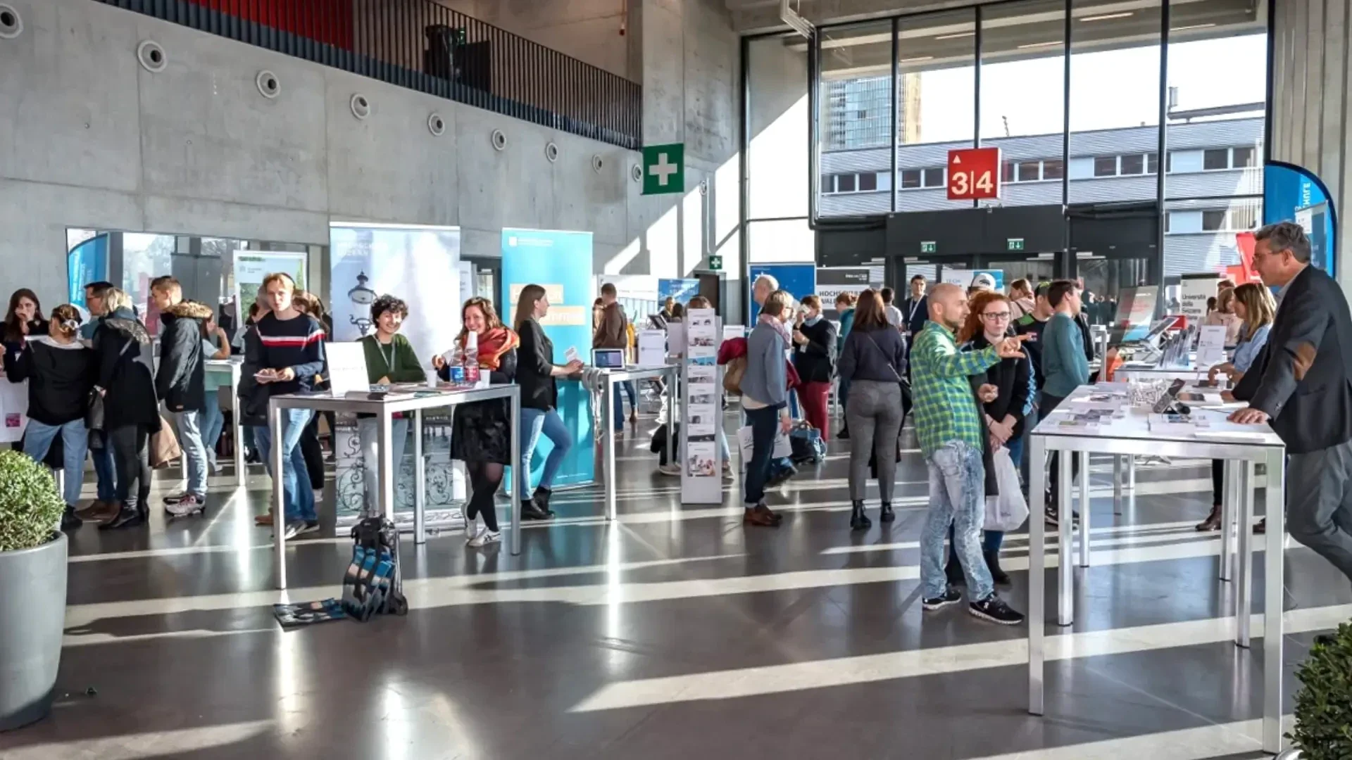 Die Menschen sind in einer hellen Halle mit Informationsständen und Tischen versammelt. Die Teilnehmer unterhalten sich, lesen Broschüren und gehen in einer geräumigen Halle mit großen Fenstern und moderner Einrichtung umher.