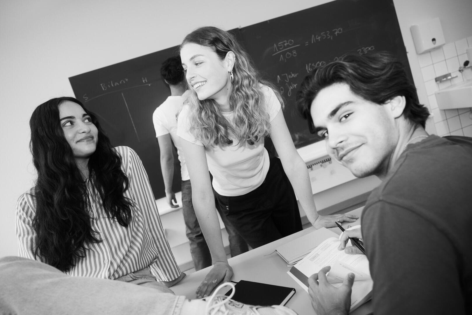 Drei Schüler unterhalten sich lächelnd in einem Klassenzimmer. Einer lehnt an einem Pult, ein anderer sitzt, und der dritte steht neben einer Tafel, auf der Notizen stehen. Die Szene wirkt ungezwungen und freundlich.