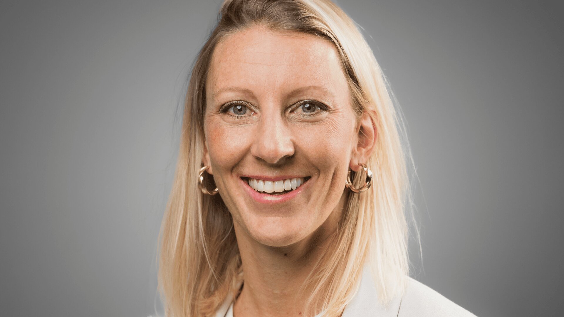 Eine Frau mit glattem blondem Haar lächelt in die Kamera. Sie trägt goldene Ohrringe und einen hellen Blazer vor einem einfarbigen grauen Hintergrund.