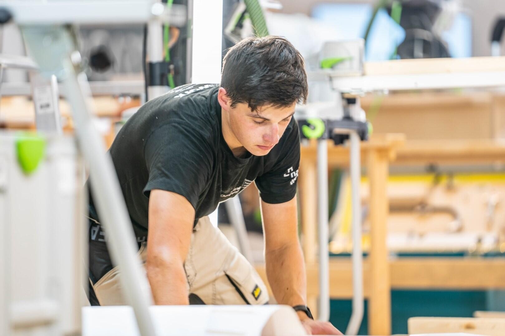 Ein junger Mann in einem schwarzen Hemd und einer beigen Hose arbeitet konzentriert in einer hellen Holzwerkstatt, über ein Stück Holz gebeugt, umgeben von Werkzeugen und Geräten.