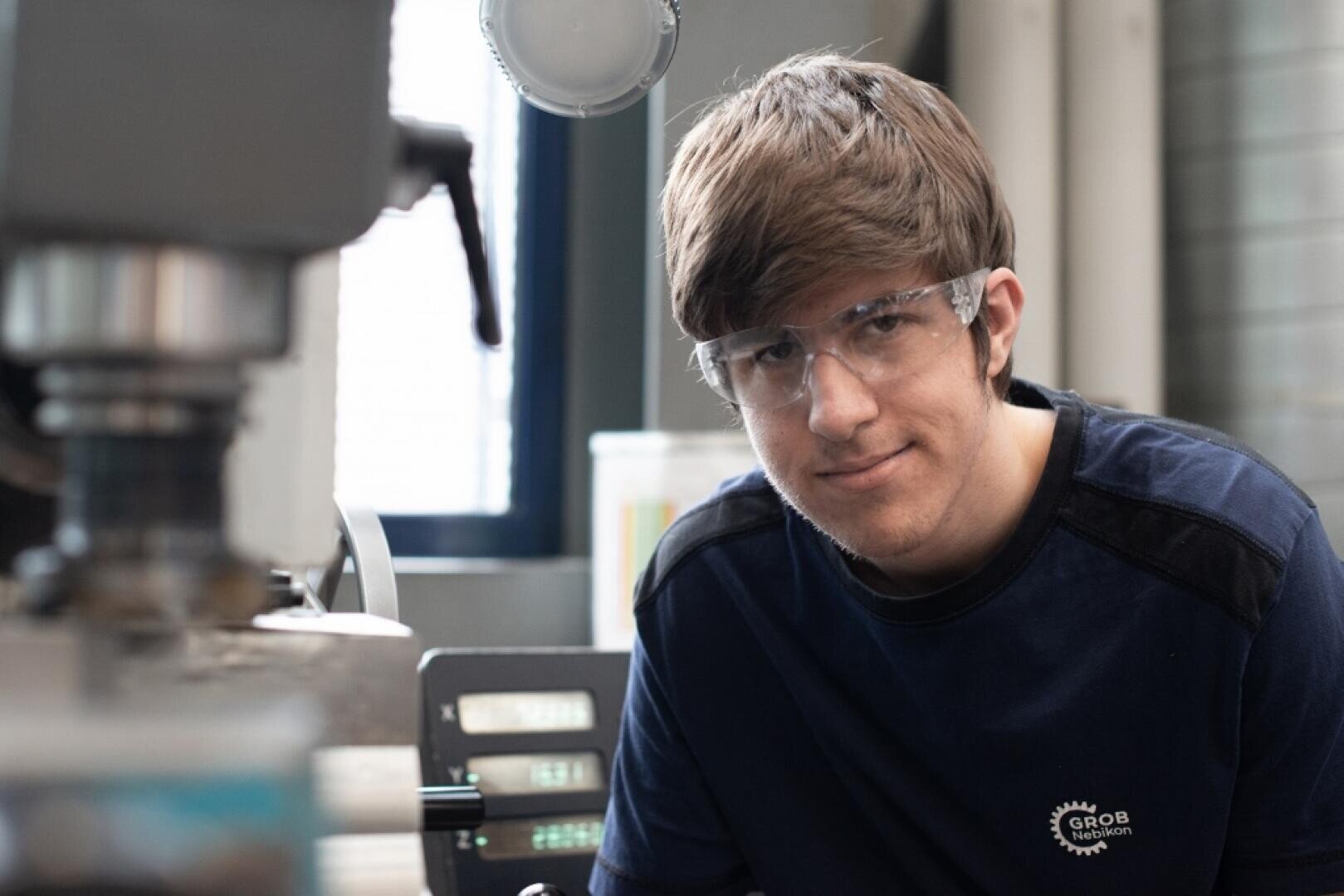 Ein junger Mann mit Schutzbrille und marineblauem Hemd steht in einer Werkstatt neben Industriemaschinen, schaut in die Kamera und lächelt leicht.