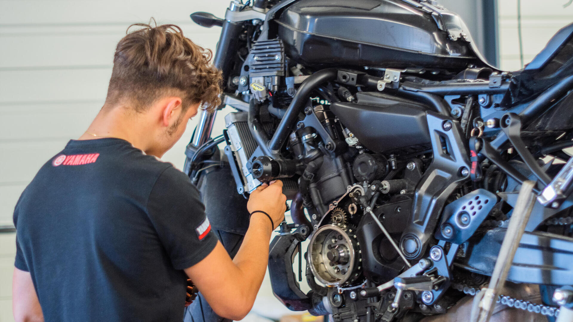 Eine Person in einem schwarzen Yamaha-T-Shirt repariert oder inspiziert den freiliegenden Motor eines schwarzen Motorrads in einer Garage oder Werkstatt.