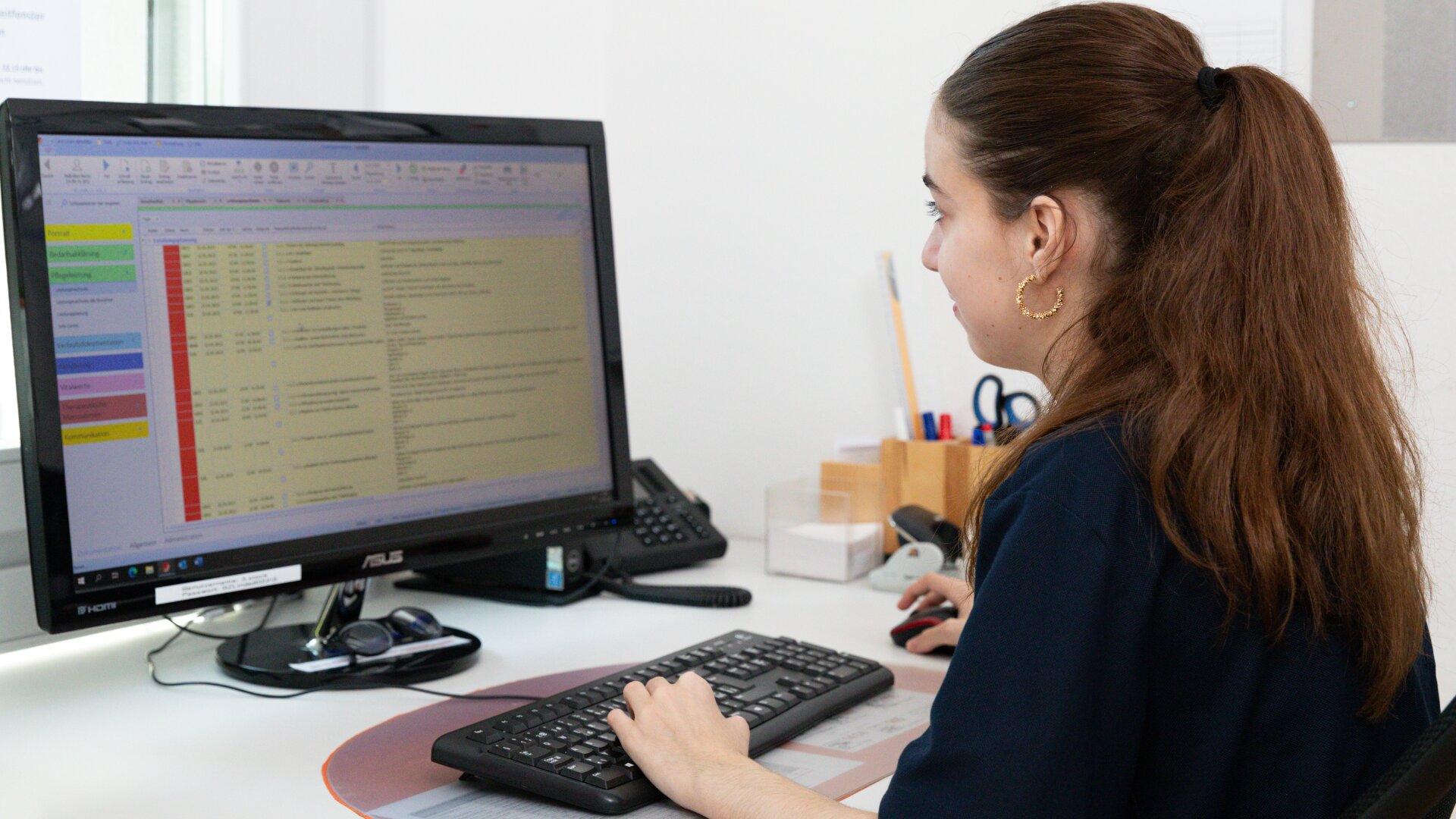 Eine Frau mit langen braunen Haaren in einem Pferdeschwanz sitzt an einem Schreibtisch und arbeitet an einem Computer mit einem großen Monitor, auf dem eine Tabellenkalkulation mit bunten Zeilen und Spalten angezeigt wird. Im Hintergrund sind Büromaterialien zu sehen.