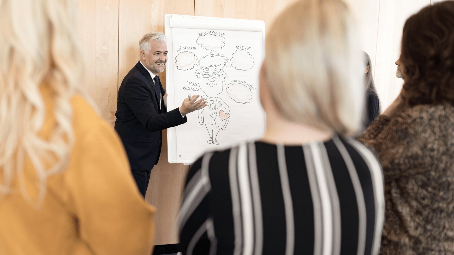 Ein Mann im Anzug präsentiert einer Gruppe von vier Personen ein Flipchart mit einer Karikaturzeichnung. Die Gruppe steht mit dem Rücken zur Kamera und hört in einem hellen Raum mit Holzwänden aufmerksam zu.