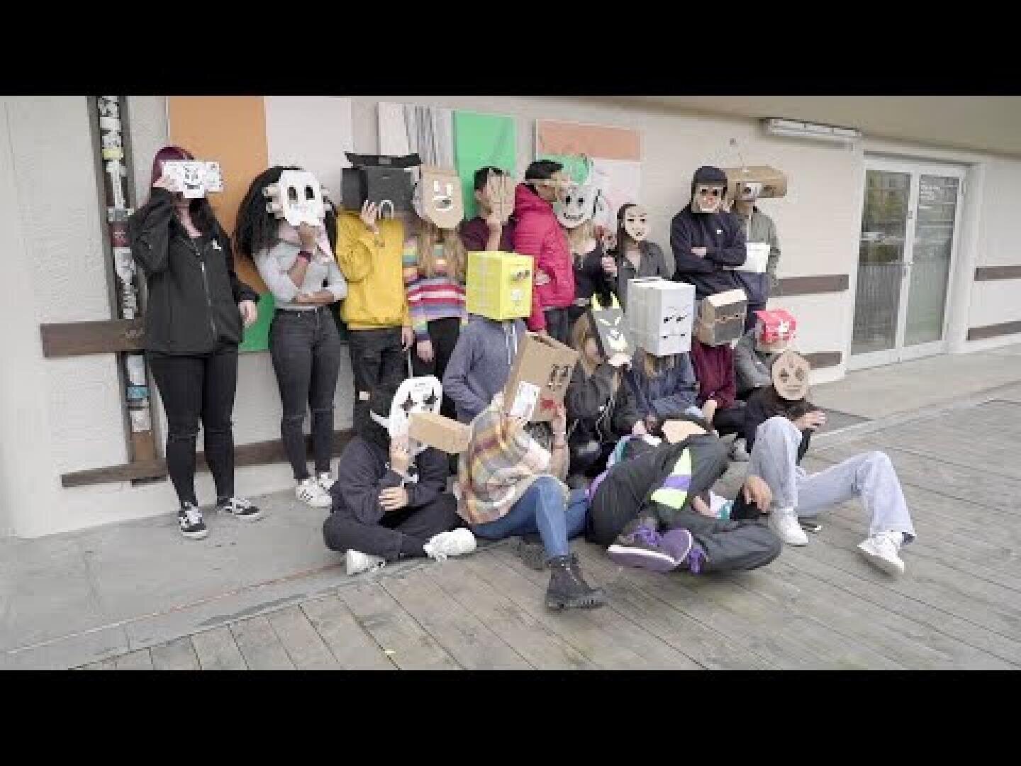 Eine Gruppe von Menschen posiert im Freien vor einem Gebäude, wobei jeder eine kreativ gestaltete Pappschachtel oder Papiermaske über dem Kopf trägt, die das Gesicht verdeckt.