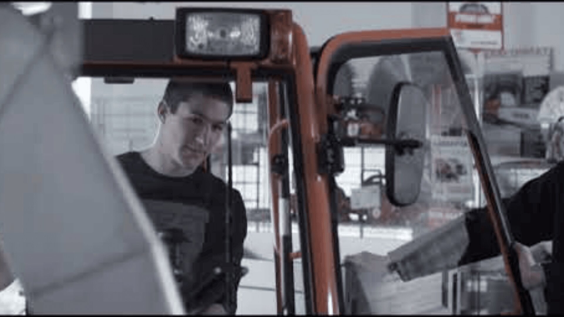 Ein Mann steht neben einer offenen Fahrzeugtür und schaut hinein. Eine andere Person, die teilweise sichtbar ist, hält ein Dokument in der Nähe der Tür. Im Hintergrund sind Regale und Fahrzeugeinrichtungen zu sehen.