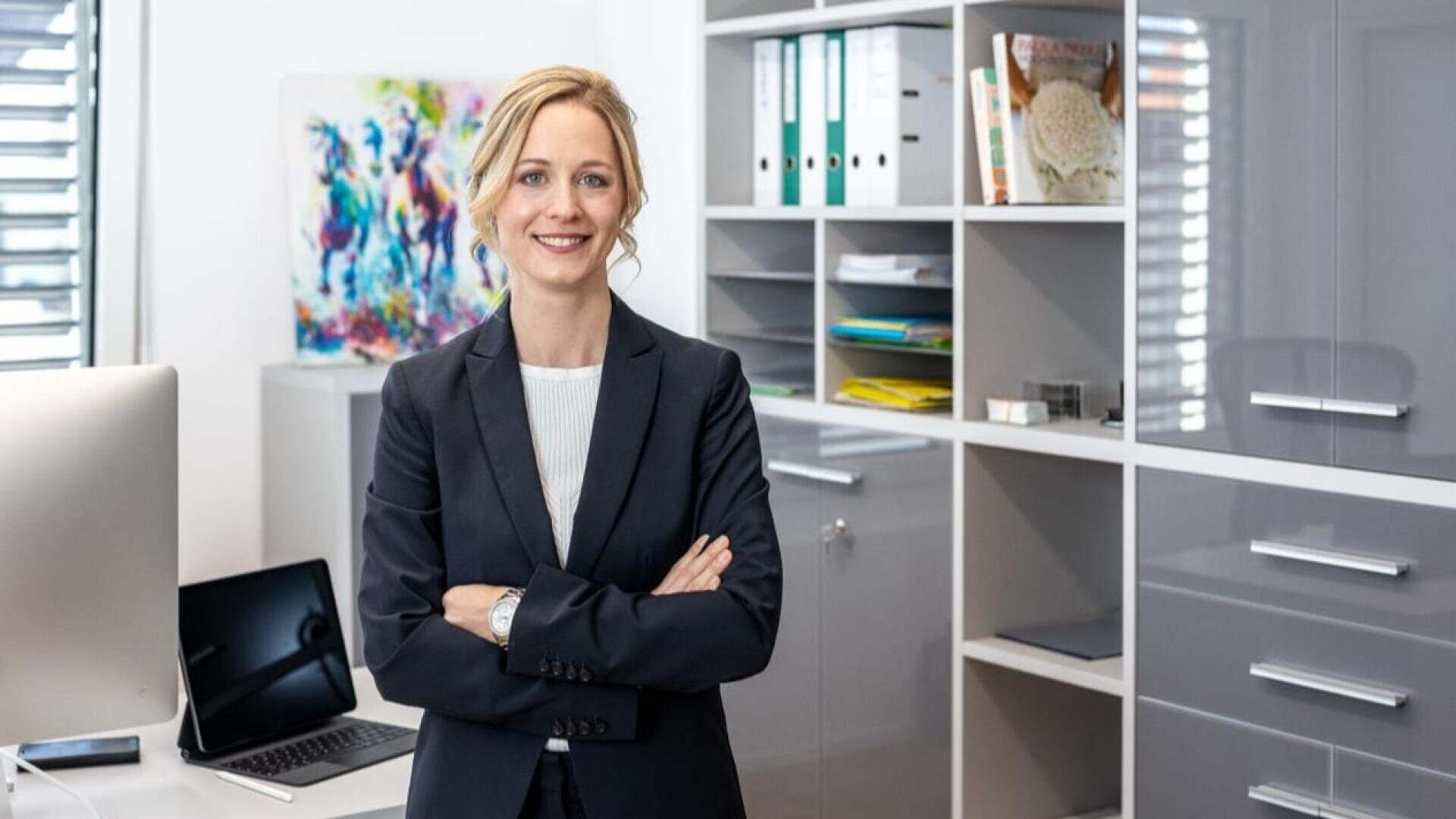 Eine Frau in einem dunklen Anzug steht lächelnd mit verschränkten Armen in einem modernen Büro, in dessen Hintergrund Regale, Aktenordner, ein Computer und Kunstwerke zu sehen sind.