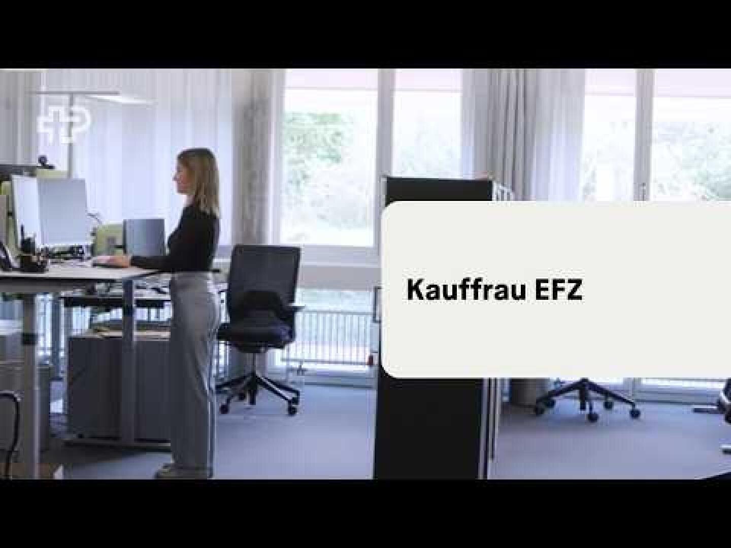 Eine Frau steht an einem höhenverstellbaren Schreibtisch in einem Büro, auf dessen rechter Seite der Schriftzug Kauffrau EFZ zu sehen ist.