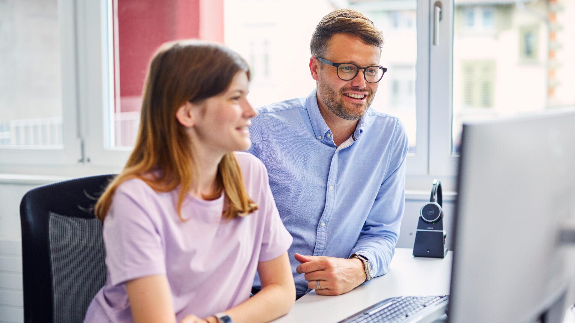 Zwei Personen sitzen nebeneinander an einem Schreibtisch vor einem Computermonitor, schauen auf den Bildschirm und lächeln. Sie befinden sich in einem hellen, modernen Büro mit großen Fenstern im Hintergrund.