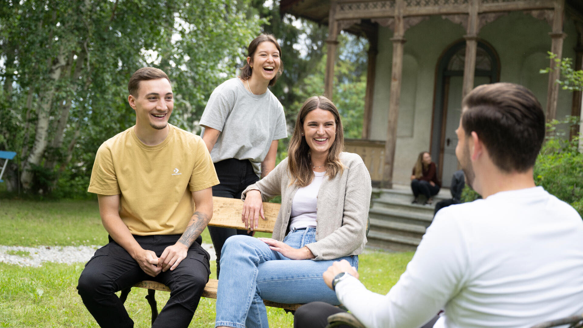 Vier junge Erwachsene sitzen und lachen gemeinsam im Freien auf einer Wiese in der Nähe eines Gebäudes mit Holzsäulen. Bäume und Grünflächen umgeben sie und schaffen eine entspannte und fröhliche Atmosphäre.