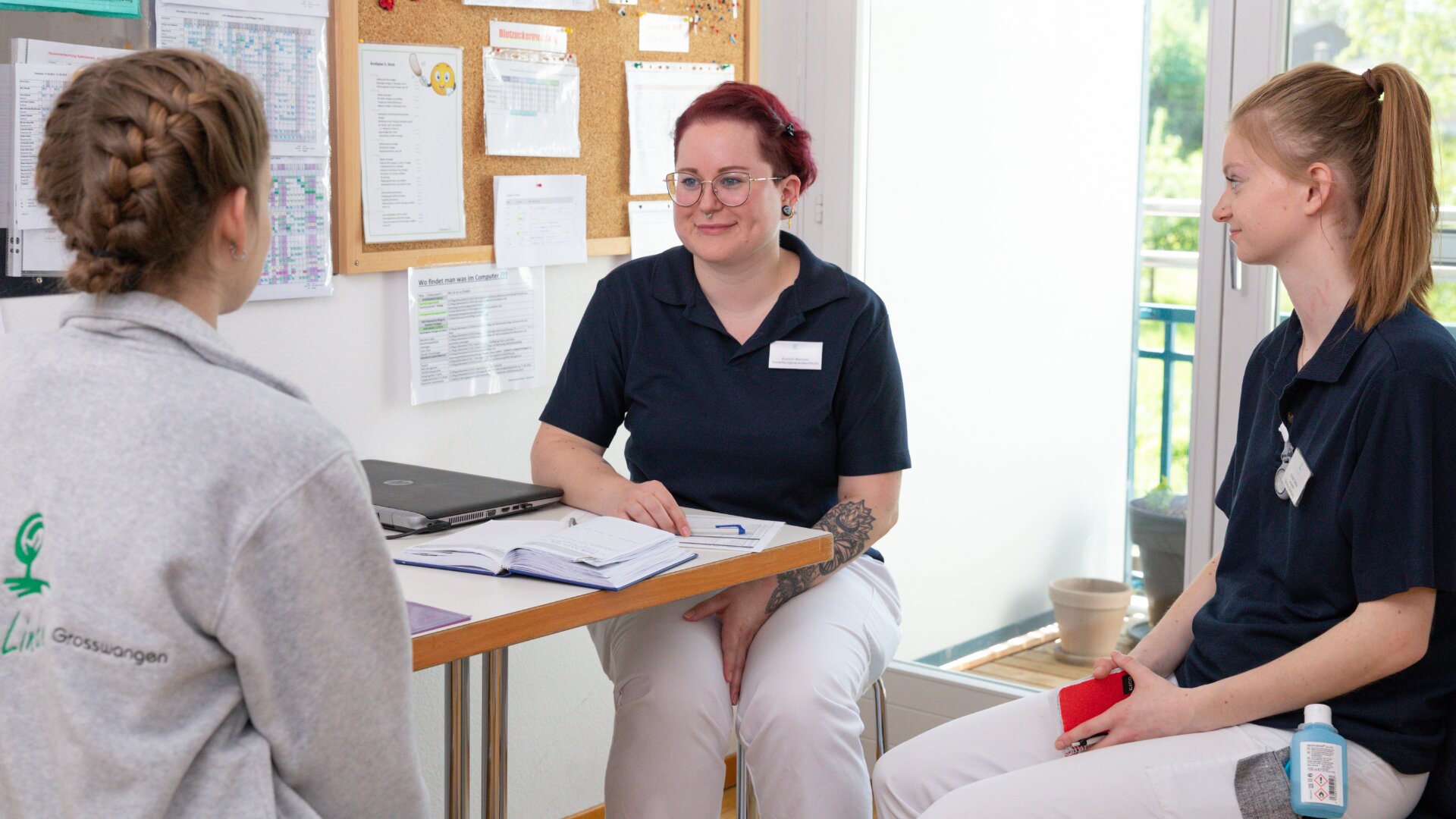 Drei Frauen führen eine Diskussion in einem Büro. Zwei Frauen in marineblauen Uniformen sitzen an einem Tisch mit Notebooks und einem Laptop, während die dritte, in einem hellgrauen Sweatshirt, ihnen gegenüber sitzt und von der Kamera wegschaut.