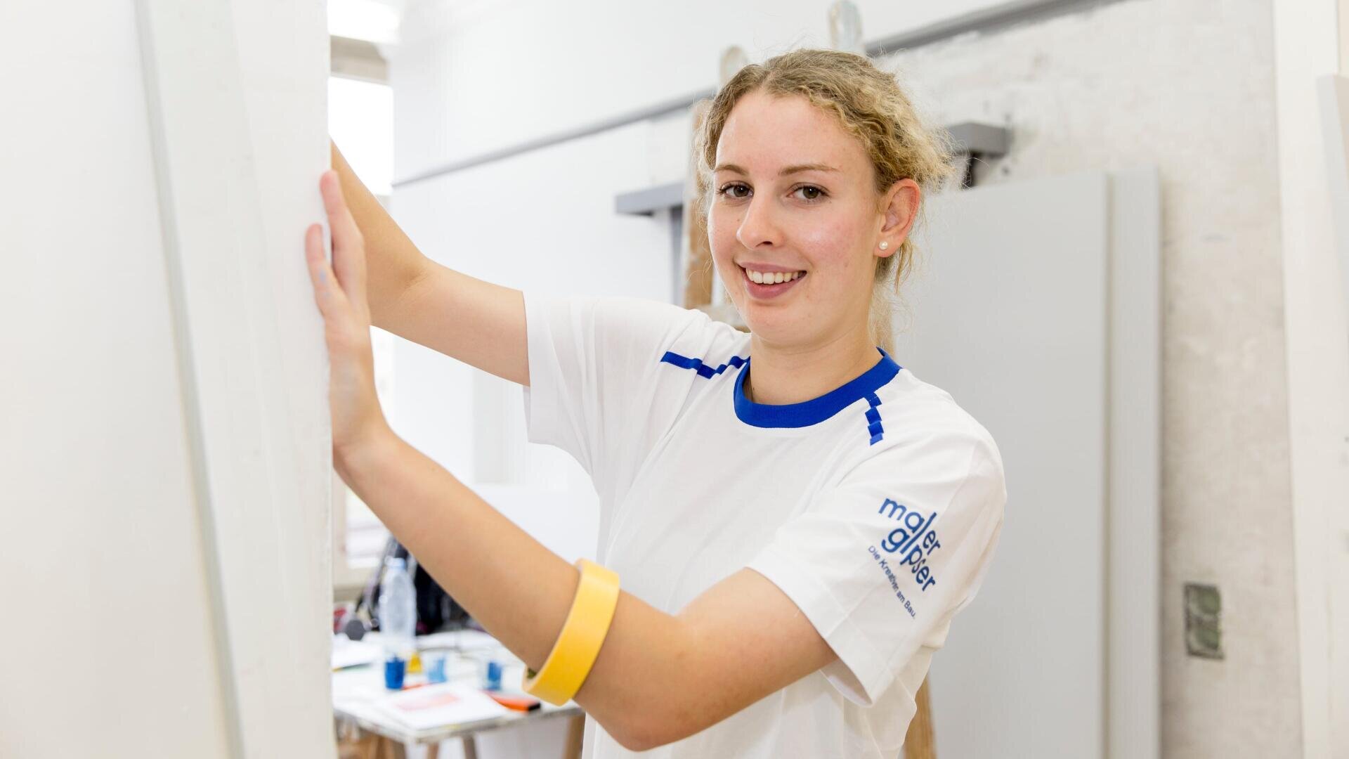 Eine lächelnde junge Frau in einem weiß-blauen T-Shirt steht in einem Haus und hält eine weiße Tafel. Im Hintergrund sind Malutensilien und andere Gegenstände unscharf zu erkennen.