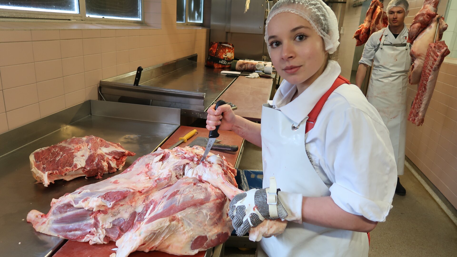 Eine Frau in Metzgerkleidung schneidet ein großes Stück rohes Fleisch auf einem Edelstahltisch in einem Fleischverarbeitungsraum, während ein anderer Metzger im Hintergrund ein Stück Fleisch in der Hand hält.