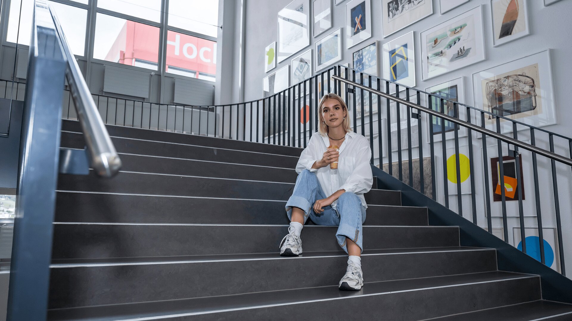 Eine junge Frau in legerer Kleidung sitzt auf einer modernen Treppe und hält eine Eiswaffel in der Hand. Sie ist umgeben von gerahmten Kunstwerken an der Wand und großen Fenstern, die eine entspannte, moderne Atmosphäre schaffen.