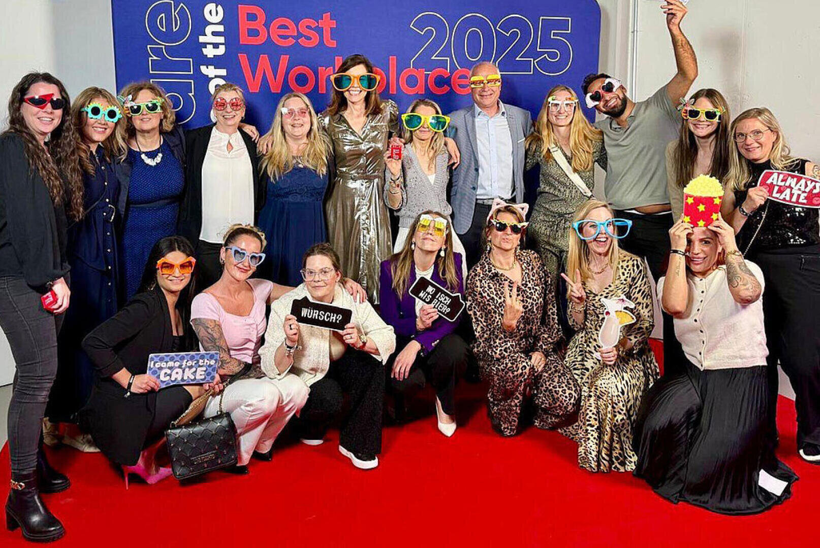 Eine Gruppe von Menschen in lustigen Outfits und mit ausgefallenen Brillen posiert gemeinsam auf einem roten Teppich vor dem Hintergrund der "Best Workplaces 2025"-Kulisse, lächelnd und mit verspielten Schildern und Requisiten.