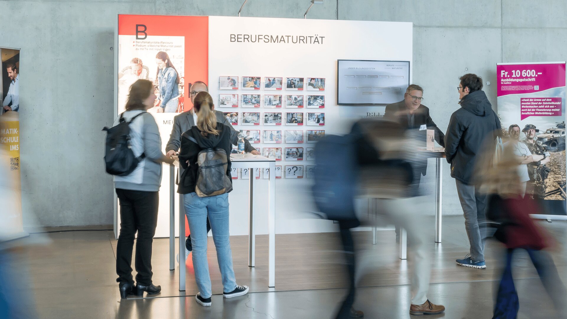 Menschen tauschen sich an einem Stand der Bildungsmesse "Berufsmaturität" aus. Mehrere Schautafeln und Informationsplakate sind zu sehen, während andere in einer hellen, modernen Halle vorbeilaufen.