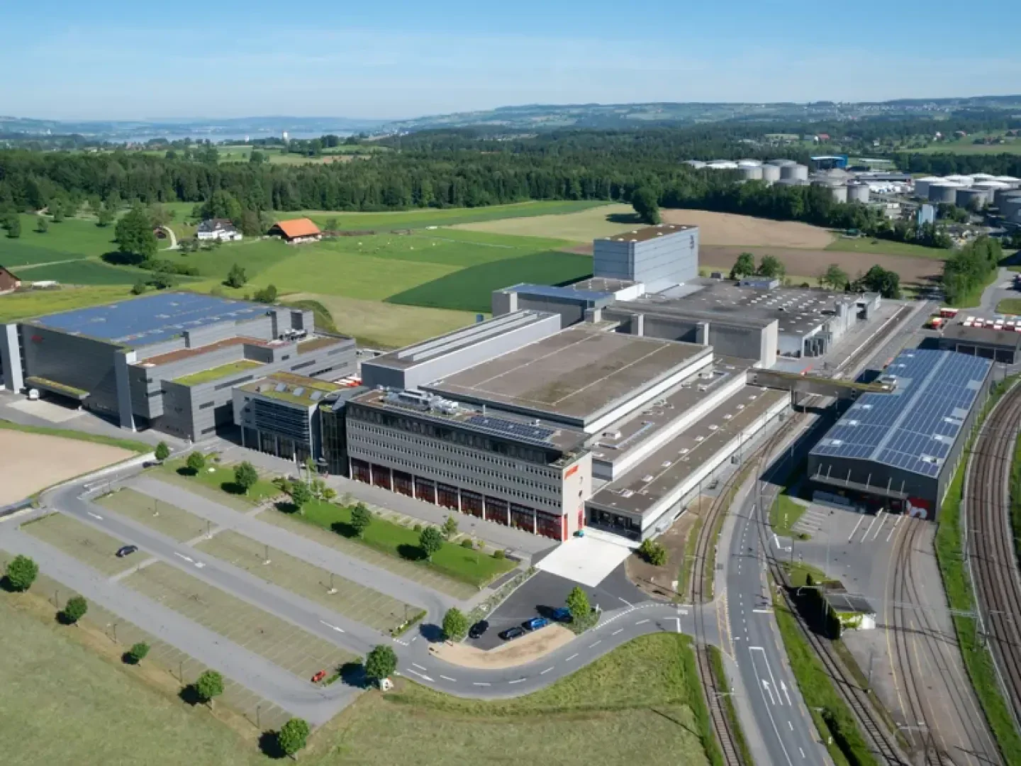 Luftaufnahme eines großen Industriekomplexes mit miteinander verbundenen Gebäuden, Parkplätzen und angrenzenden Bahngleisen in einer ländlichen Landschaft.