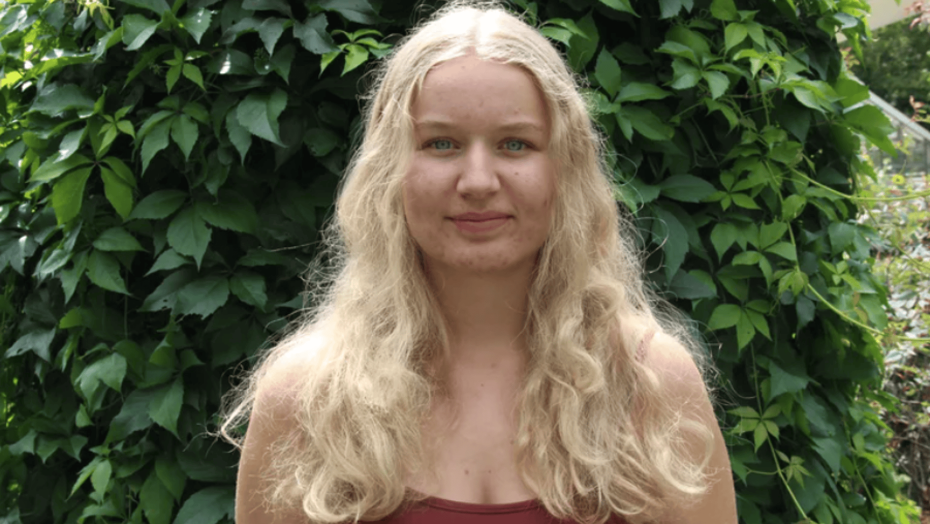 Eine junge Frau mit langen, gewellten blonden Haaren und einem kastanienbraunen Tanktop steht im Freien vor üppigem Grün und lächelt sanft in die Kamera.