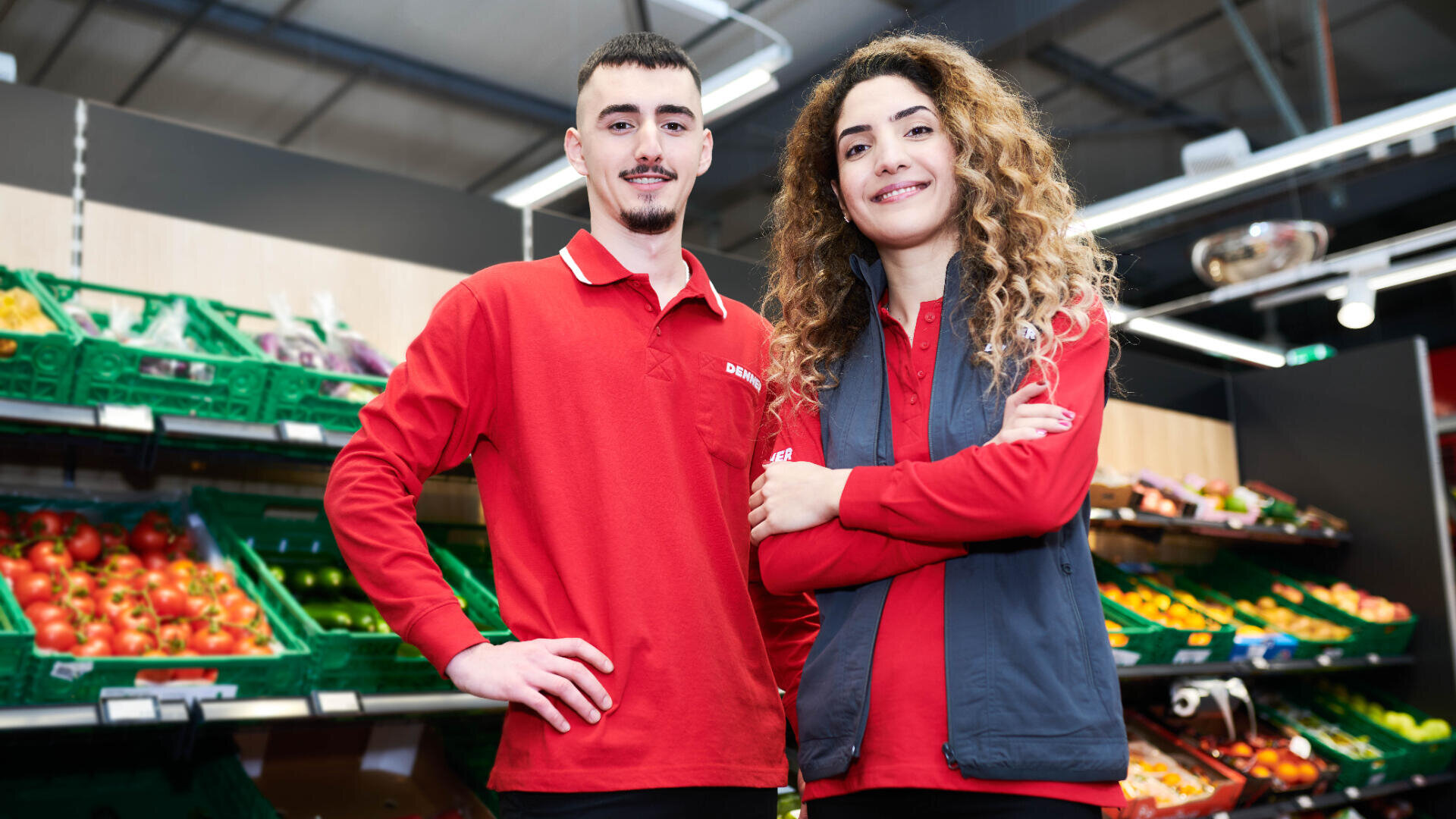 Zwei Supermarktangestellte in roten Uniformen stehen lächelnd in der Obst- und Gemüseabteilung, während hinter ihnen frisches Obst und Gemüse in den Regalen liegt.