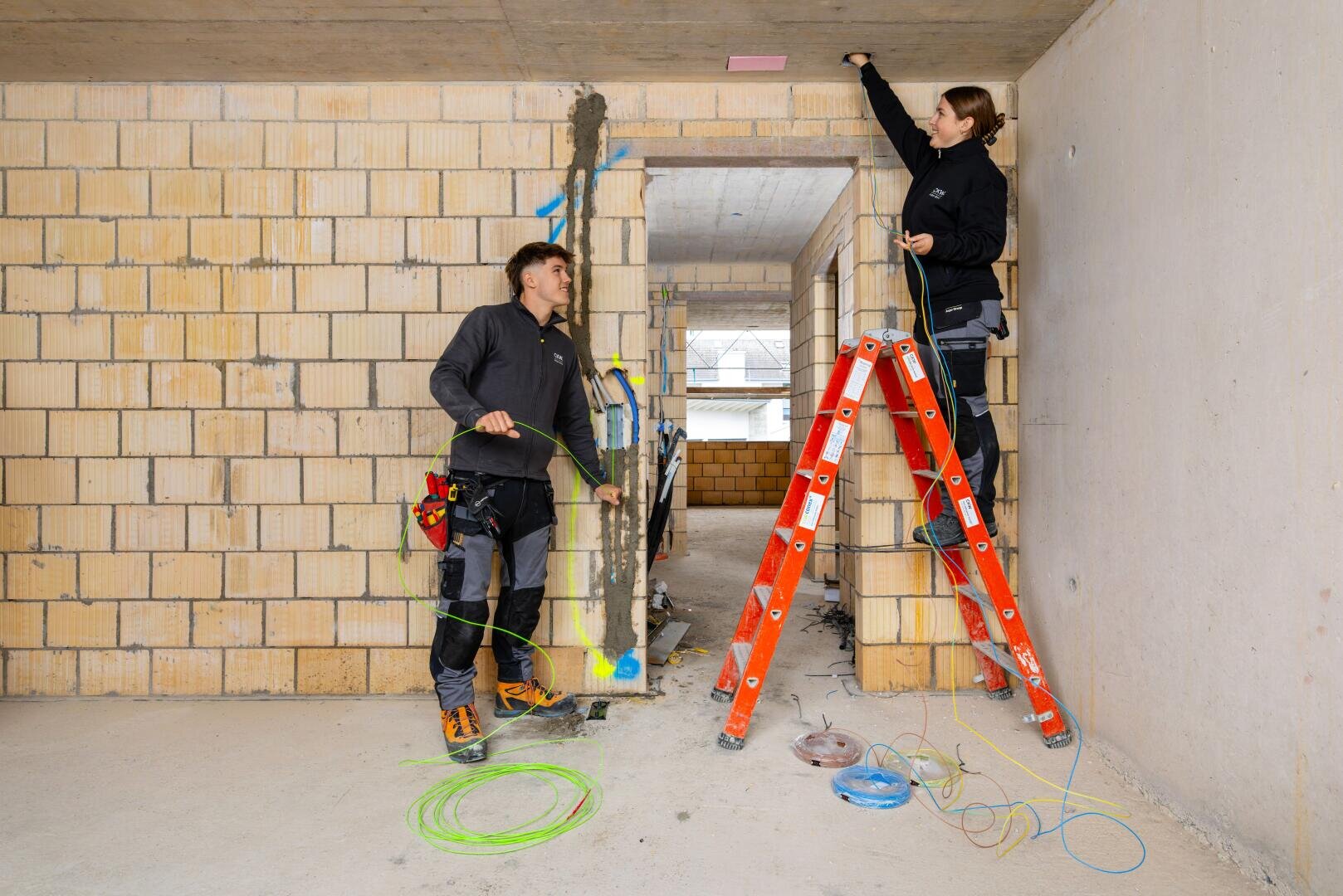 Zwei Elektriker arbeiten in einem im Bau befindlichen Gebäude. Der eine steht auf einer Leiter und befestigt ein Kabel an der Decke, während der andere auf dem Boden steht und Werkzeuge und Kabel hält. Um sie herum liegen aufgerollte Kabel und Werkzeuge verstreut.