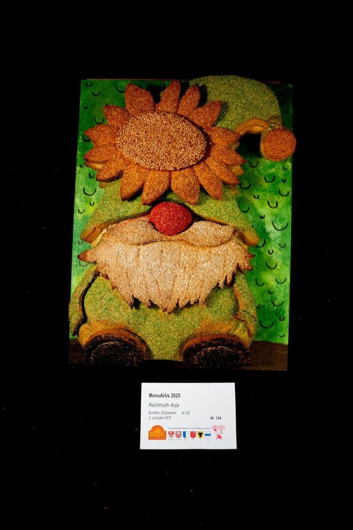 Auf grünem Hintergrund ist ein strukturiertes Kunstwerk in Form eines Gnoms mit Sonnenblumenhut und weißem Bart abgebildet. Unterhalb des Kunstwerks befindet sich ein weißes Etikett mit Textangaben. Der Hintergrund ist schwarz.