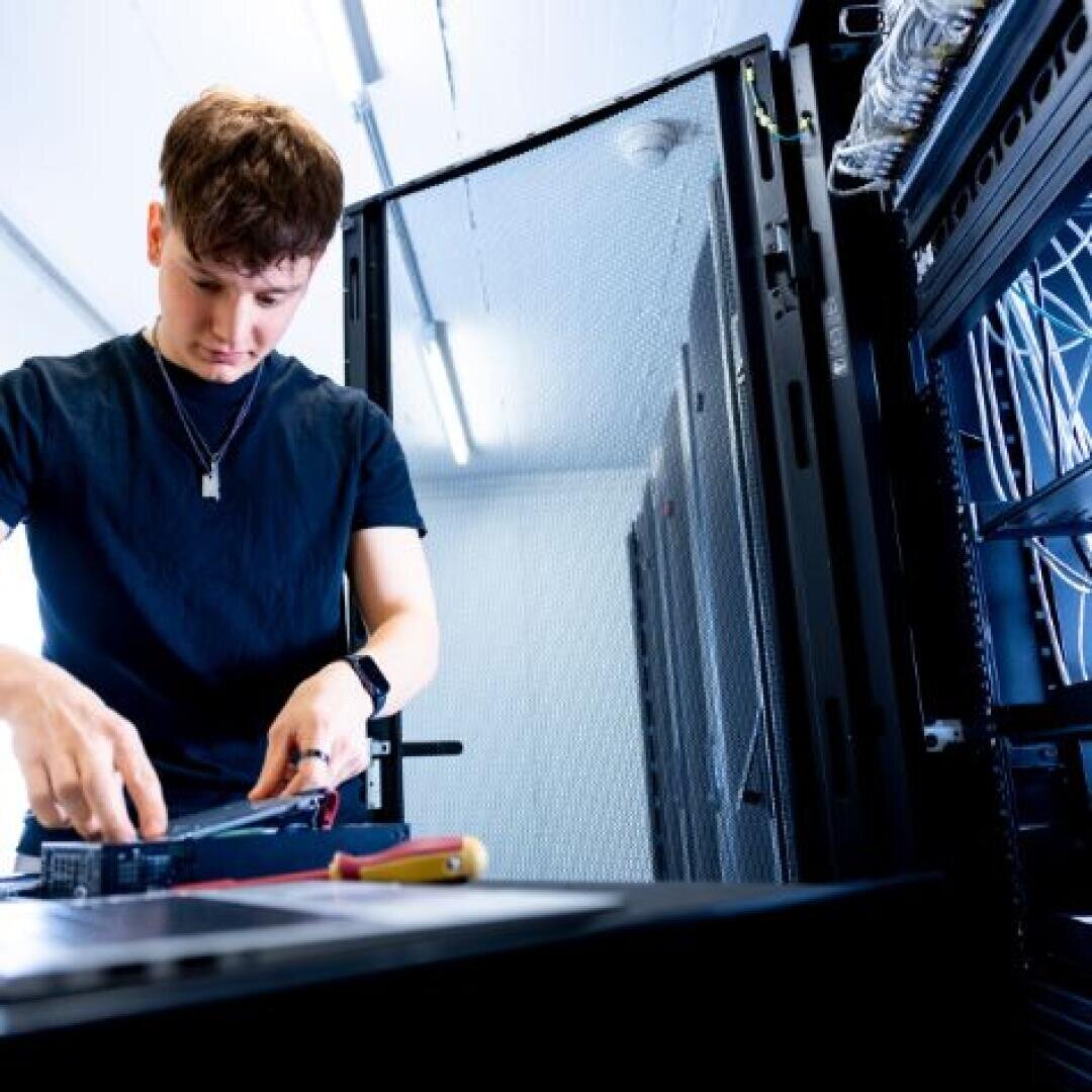 Eine Person in einem schwarzen Hemd arbeitet an elektronischen Geräten in einem Serverraum und benutzt Werkzeuge an einem Serverschrank, um den herum Kabel und offene Schränke zu sehen sind.