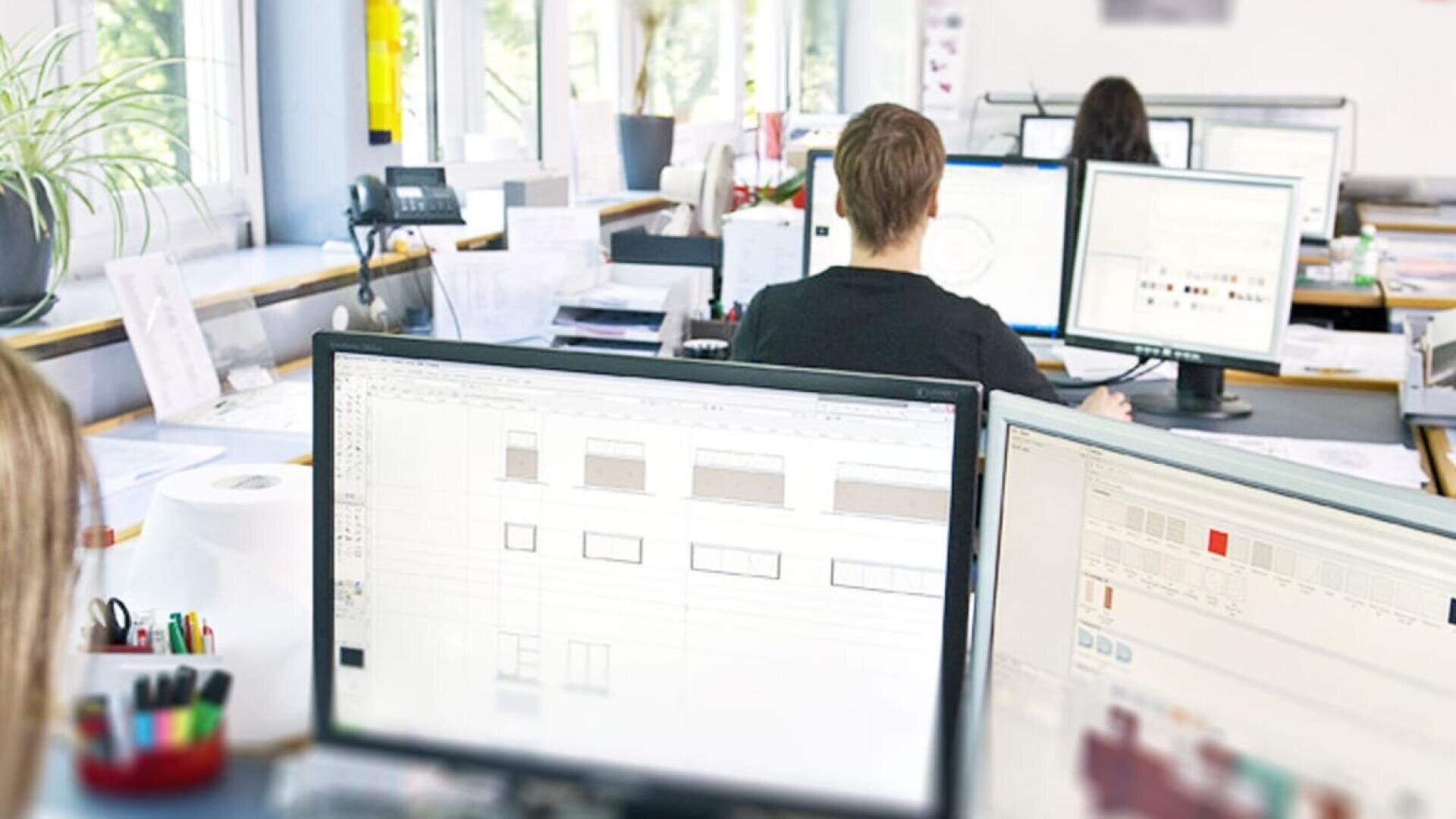 Mehrere Personen arbeiten an Schreibtischen mit Computermonitoren, auf denen Architektur- oder Designsoftware angezeigt wird, in einem hellen, modernen Büro mit großen Fenstern und verstreutem Papierkram.
