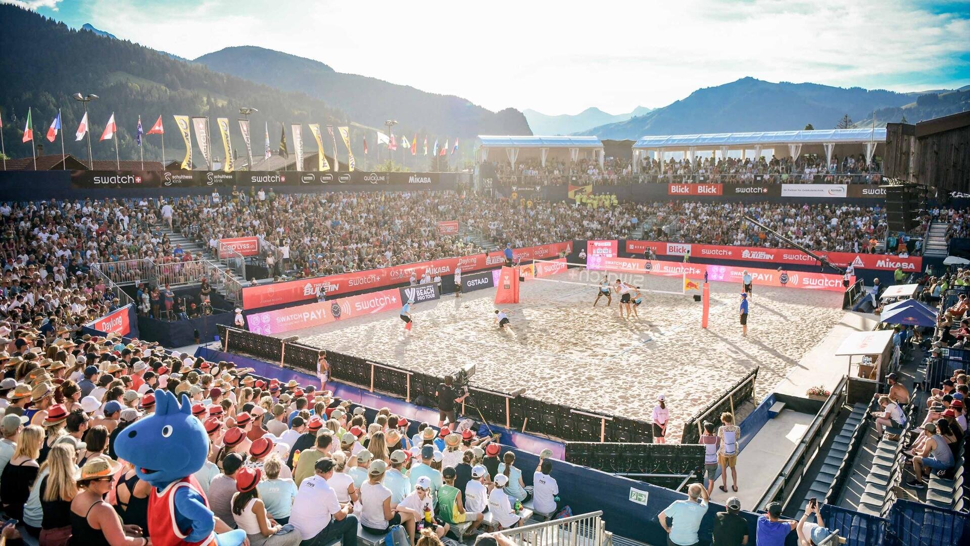 Ein volles Stadion mit jubelnden Fans umgibt ein sandiges Beachvolleyballfeld vor der Kulisse der Berge unter einem strahlenden Himmel. Die Spieler sind auf dem Platz, und ein blaues Maskottchen steht inmitten der Menge.