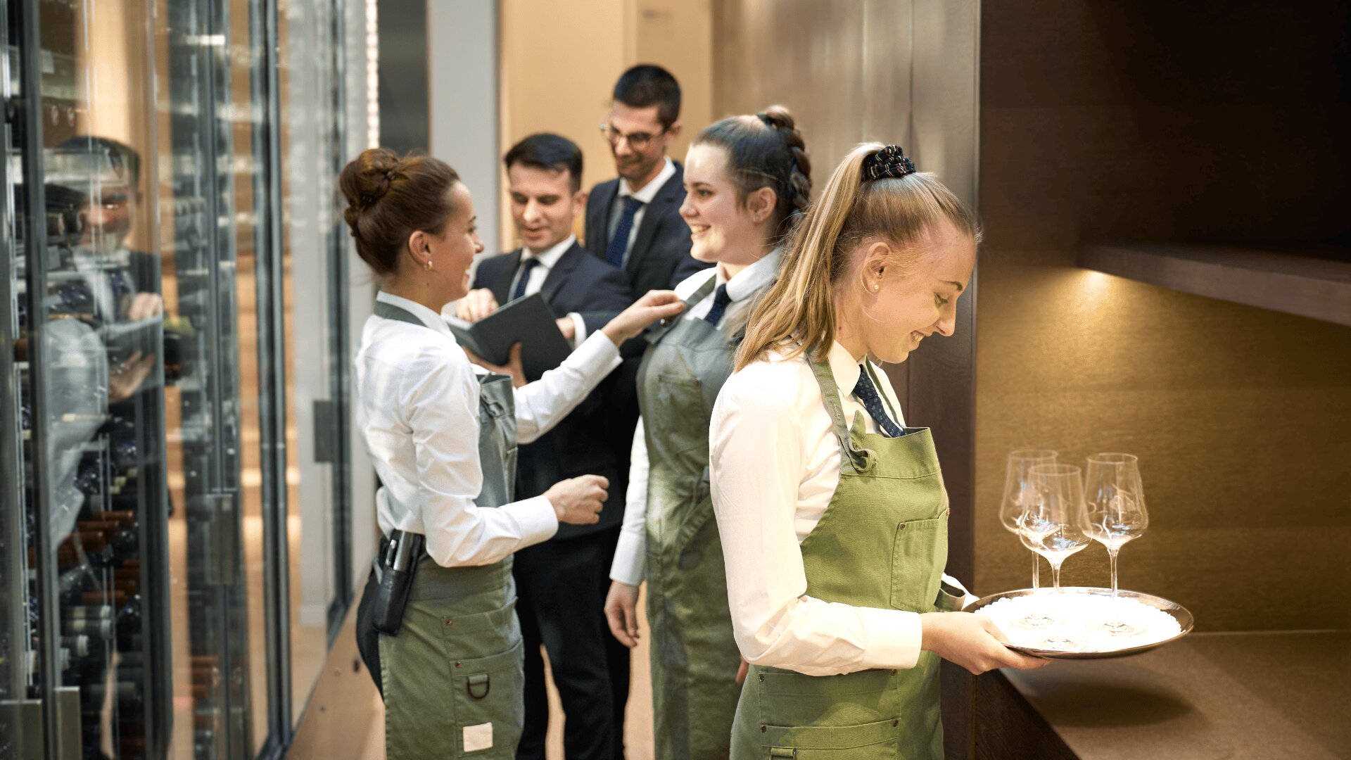 Eine Gruppe von Kellnern in grünen Schürzen und weißen Hemden bereitet sich auf den Service vor, passt ihre Uniformen an und trägt ein Tablett mit Weingläsern, während zwei Manager in einem modernen Restaurant die Aufsicht führen.