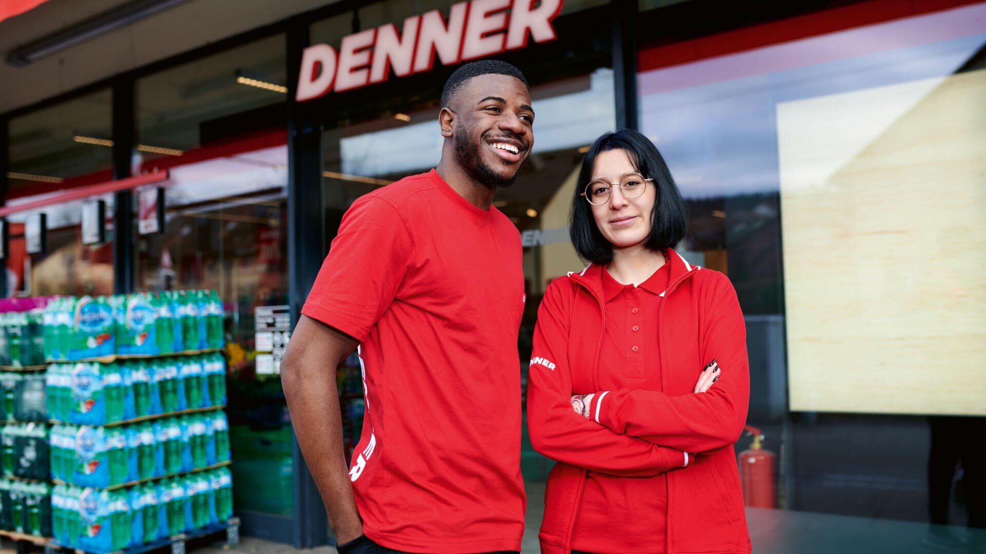 Zwei Angestellte in roten Uniformen stehen lächelnd und posierend vor einem Denner-Supermarkt, vor dessen Eingang sich Getränkeflaschen stapeln.