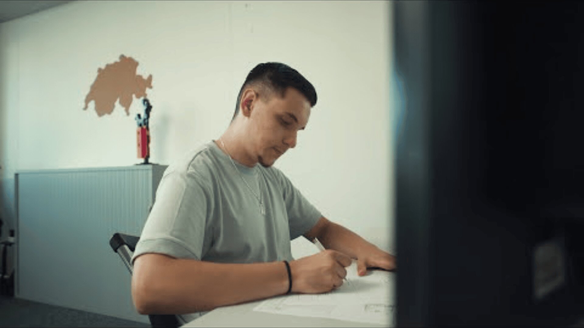 Ein junger Mann sitzt an einem Schreibtisch und schreibt in einem gut beleuchteten Raum auf Papier. An der Wand hinter ihm hängt eine Landkarte der Schweiz, und auf einem Regal steht eine rote Figur.